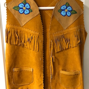 Op de afbeelding: Een bruine suède vest met franjes en kralenbloem accenten. De vest heeft twee zakken en een strik sluiting.