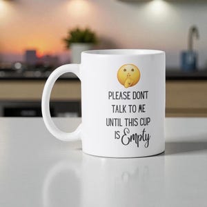 May include: White ceramic mug with a handle, featuring the text "PLEASE DONT TALK TO ME UNTIL THIS CUP IS Empty" in black script. An emoji of a face with a finger over its lips is above the text. The mug is on a white surface.