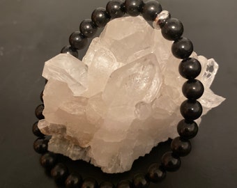 May include: A black beaded bracelet with a silver bead accent. The bracelet is resting on a cluster of white crystal rocks.