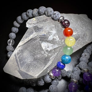 May include: A black lava rock bracelet with silver beads and a rainbow of chakra stones. The bracelet is resting on a large clear quartz crystal.