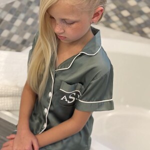 Op de afbeelding: Een jong persoon draagt een groene satijnen pyjamaset met witte piping. De pyjama heeft een zakje aan de linkerkant met de initialen "AS" in wit geborduurd.