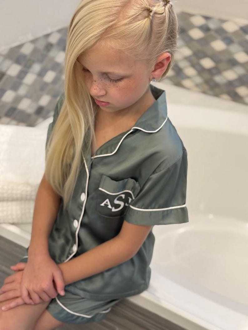 Op de afbeelding: Een jong persoon draagt een groene satijnen pyjamaset met witte piping. De pyjama heeft een zakje aan de linkerkant met de initialen "AS" in wit geborduurd.