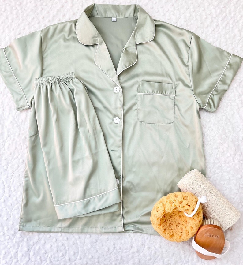 May include: A sage green satin pajama set with short sleeves and a button-up top. The set includes a top with a collar and a pocket, and matching shorts.