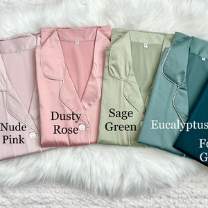 May include: Six satin pajamas in different colors: blush, nude pink, dusty rose, sage green, eucalyptus, and forest green. Each pajama top has a button closure and a collar.