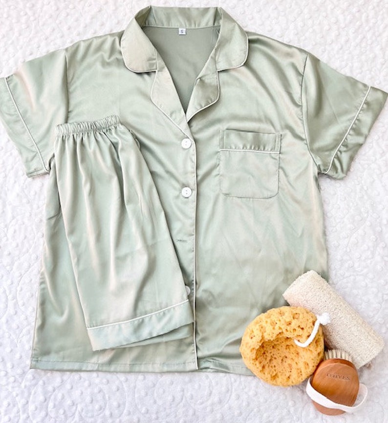 Op de afbeelding: Een groene satijnen pyjamaset met korte mouwen en een knoopsluiting. Het bovenstuk heeft een zak aan de linkerkant en een witte rand rond de kraag en mouwen. De broek heeft een witte rand rond de pijpen.