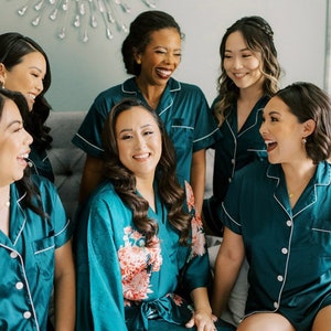 Op de afbeelding: Vijf vrouwen in tealblauwe pyjama's en badjassen. De vrouw in het midden draagt een tealblauwe badjas met een bloemenprint. De vrouwen lachen en glimlachen.