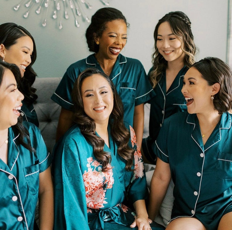 Op de afbeelding: Vijf vrouwen in tealblauwe pyjama's en badjassen. De vrouw in het midden draagt een tealblauwe badjas met een bloemenprint. De vrouwen lachen en glimlachen.