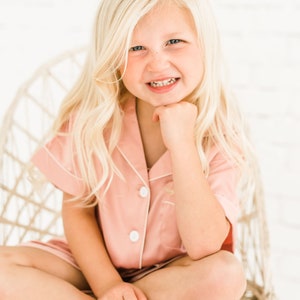 May include: A young girl with blonde hair is sitting in a wicker chair. She is wearing a pink pajama top with white piping and white buttons. She is smiling and has her hand on her chin.