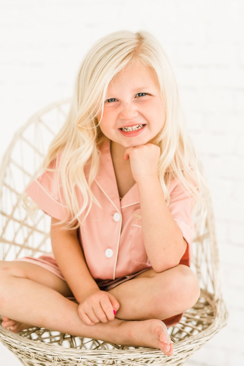 May include: A young girl with blonde hair is sitting in a wicker chair. She is wearing a pink pajama top with white piping and white buttons. She is smiling and has her hand on her chin.