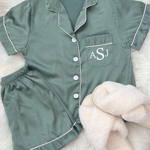 May include: A sage green satin pajama set with short sleeves and shorts. The top has a white button closure and a white pocket with the initials ASJ embroidered on it. The set is paired with white fluffy slippers.