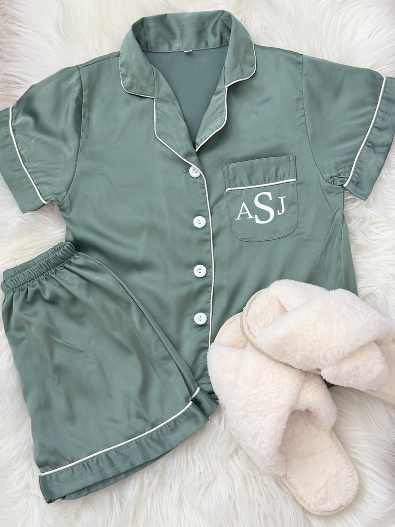May include: A sage green satin pajama set with short sleeves and shorts. The top has a white button closure and a white pocket with the initials ASJ embroidered on it. The set is paired with white fluffy slippers.