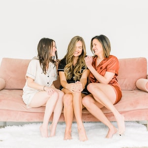 Op de afbeelding: Drie vrouwen in pyjama zitten op een roze fluwelen bank. Ze lachen en praten met elkaar. De bank staat in een witte kamer met een wit tapijt.
