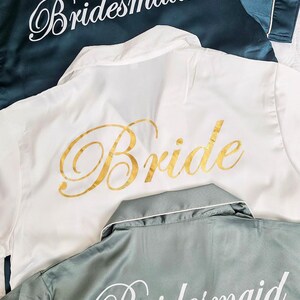 Op de afbeelding: Drie satijnen gewaden met de woorden "Bride" en "Bridesmaid" geborduurd. De gewaden zijn in wit, teal en saliegroen.