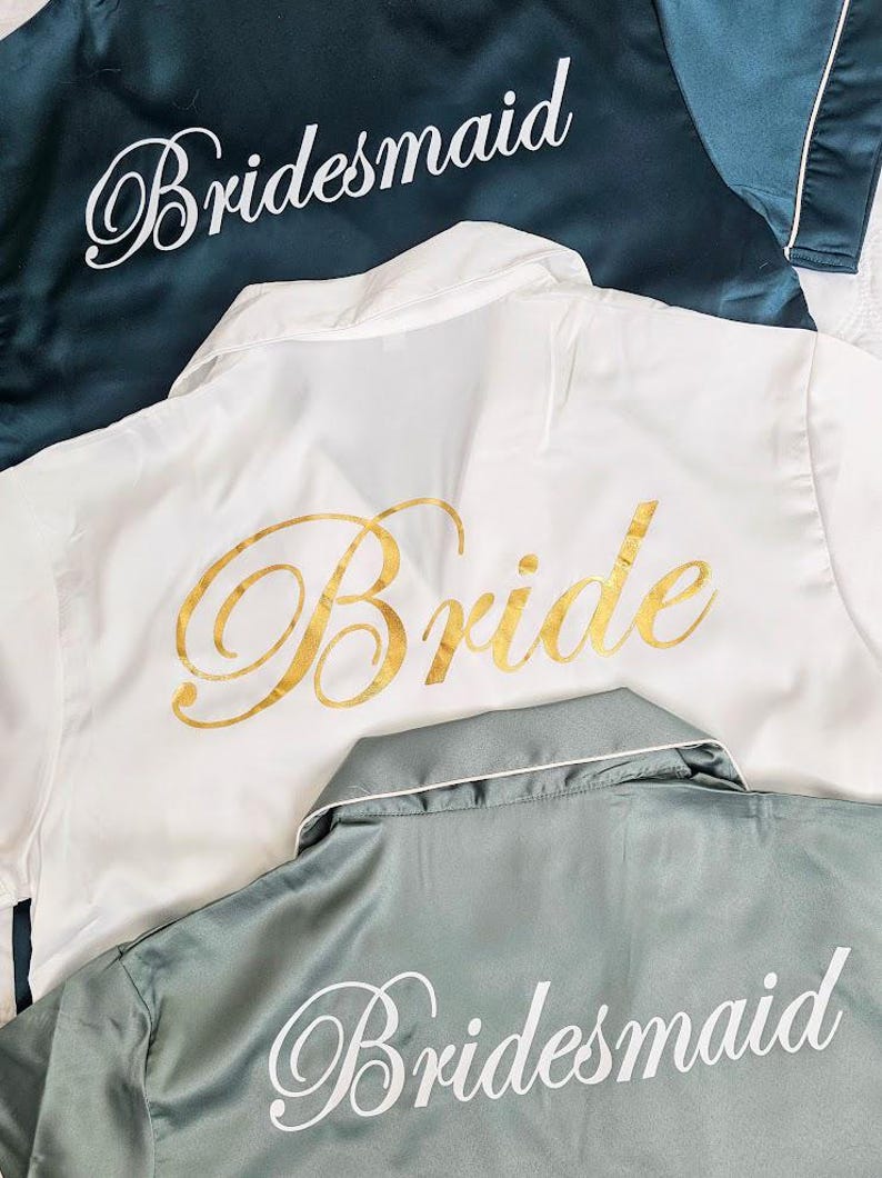 Op de afbeelding: Drie satijnen gewaden met de woorden "Bride" en "Bridesmaid" geborduurd. De gewaden zijn in wit, teal en saliegroen.