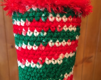 May include: A handmade, crocheted cup cozy in red, green, and white. The cozy has a striped pattern and a fluffy red trim at the top. The cozy is designed to fit a standard-sized disposable cup. The cozy is a festive accessory.