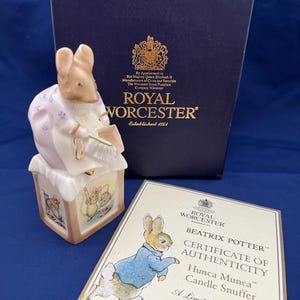 Puede incluir: Una figurita de porcelana blanca de un ratón sosteniendo un libro, sentado en una pequeña casa con un techo marrón. La figurita es parte de una colección de Beatrix Potter y es una edición limitada de 500. La caja es azul con letras doradas y el certificado de autenticidad es blanco con letras doradas.