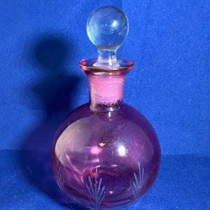 Puede incluir: Un tapón de botella redondo de vidrio transparente con una botella de vidrio rosa con diseños grabados. La botella es de color rosa intenso.