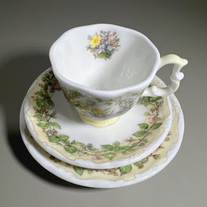 Puede incluir: Un juego de té vintage que incluye una taza, un platillo y un plato pequeño. La taza de porcelana blanca presenta un diseño floral y un asa amarilla. El platillo y el plato pequeño tienen un patrón floral y de hojas a juego con un borde amarillo. Un juego de té clásico.