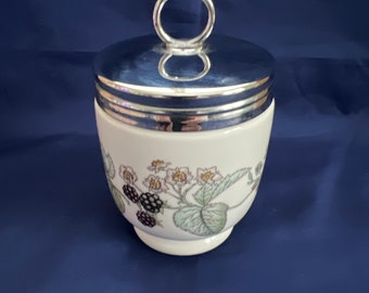 Könnte beinhalten: Eine weiße Keramik-Eierbecher mit einem silbernen Deckel und einem Blumendesign mit Brombeeren und weißen Blüten. Der Deckel hat einen silbernen Ringgriff.