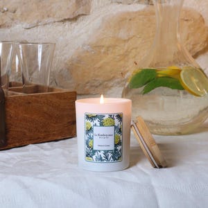 Könnte beinhalten: Eine brennende weiße Kerze mit floralem Etikett und Holzdeckel steht auf einer weißen Tischdecke. Auf dem Etikett steht "La Flamboyante Bougies" und "Maquis Corse". Im Hintergrund ein Glaskrug mit Zitronenscheiben und Minze.