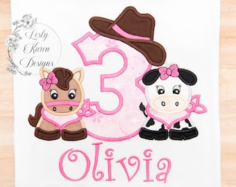May include: A white t-shirt featuring an embroidered design of a brown horse and a black and white cow, both wearing pink bows and bandanas. A large pink number three with a brown cowboy hat sits between them, with the name Olivia in pink.