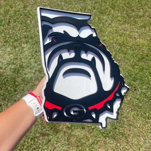 May include: A layered wooden sign in the shape of the state of Georgia, featuring the University of Georgia Bulldogs mascot. The sign is black, white, and red, with the letter "G" visible.