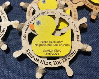 May include: Wooden wheel-shaped duck toy with a yellow duck in the center. The wheel has the text "CON*QUACK*ULATIONS you found a cruising duck! KEEP OR HIDE, YOU DECIDE!" and a yellow tag that says "Public places only. Carnival Glory 8/16/2024".