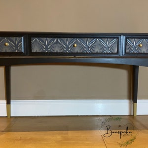 May include: A black console table with three drawers featuring a fan-shaped design in silver and grey. The drawers have gold knobs. The table has gold-coloured feet and a curved leg design. The table is against a neutral-coloured wall.