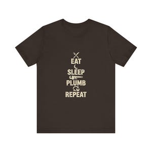 May include: A dark brown short-sleeve t-shirt featuring a graphic print in a distressed beige font. The design displays the text EAT SLEEP PLUMB REPEAT, with a fork and knife icon above EAT, a crescent moon above SLEEP, a pipe wrench above PLUMB, and arrows above REPEAT.