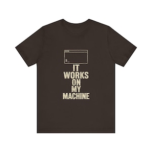 Peut inclure: Un t-shirt à manches courtes marron foncé avec un graphique blanc représentant une icône de fenêtre de terminal informatique au-dessus du texte "IT WORKS ON MY MACHINE" dans une police de style bloc vieillie centrée sur la poitrine.