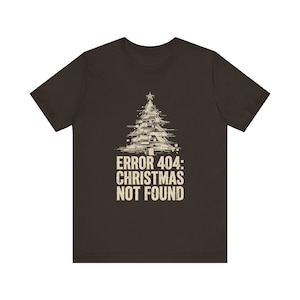 Peut inclure: Un t-shirt à manches courtes marron foncé avec un motif de sapin de Noël stylisé à effet glitch surmonté d'une étoile. Sous l'arbre, le texte "ERROR 404: CHRISTMAS NOT FOUND" est imprimé dans une police sans empattement claire et usée.