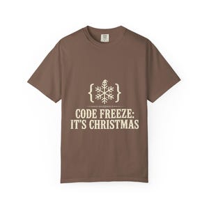 May include: A brown short-sleeved t-shirt featuring a white graphic of a snowflake inside curly brackets with a small Santa hat on top. Below the graphic, the text reads CODE FREEZE: IT'S CHRISTMAS in a distressed, off-white, capitalized font.