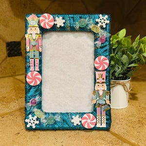 May include: A teal picture frame decorated with candy, snowflakes, and nutcracker figures. The frame has a white center and is adorned with pink and white peppermint candies, clear candies, and snowflake accents. The nutcrackers are in pink, teal, and gold.