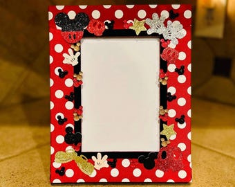 May include: Red picture frame with white polka dots and Mickey Mouse themed decorations. The frame has a black border and a white mat. The decorations include Mickey Mouse ears, gloves, and stars.