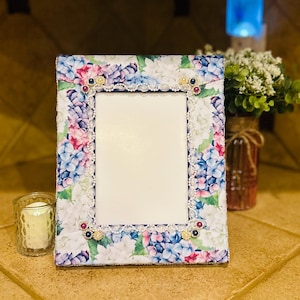 May include: A rectangular picture frame with a floral hydrangea pattern in shades of blue, pink, and white. The frame is embellished with lace trim and decorative jewels. A candle in a glass votive sits to the left.