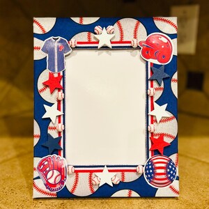 May include: A baseball-themed picture frame with a blue background patterned with baseballs. The frame is decorated with baseball-related embellishments, including a jersey, helmet, glove, stars, and an American flag baseball. The frame is approximately 8x10 inches.