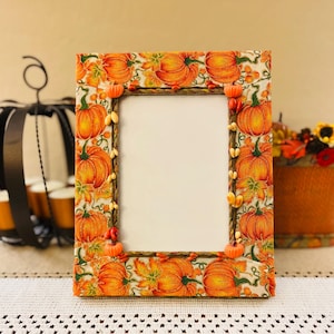 May include: A rectangular picture frame featuring a fabric cover with a vibrant orange pumpkin and autumn leaf pattern. The inner border is accented with a twisted rope trim, small decorative orange pumpkin beads, and scattered light-colored seeds.