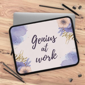 May include: A laptop sleeve with a black border and a light background. The sleeve features the text "Genius at work" in dark purple script, along with watercolor floral accents in shades of purple, peach, and gold.