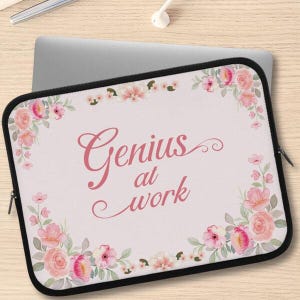 May include: A laptop sleeve with a black zipper and trim. The sleeve has a light pink background with the words "Genius at work" in a cursive font. Pink and peach floral designs decorate the corners.