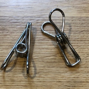 May include: Two silver metal clothespins with a spring in the middle. The clothespins are on a wooden surface.