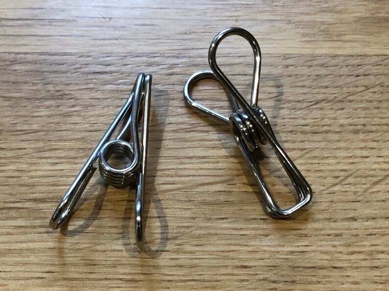 May include: Two silver metal clothespins with a spring in the middle. The clothespins are on a wooden surface.