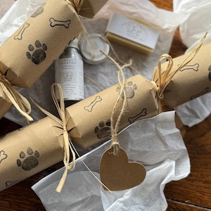 Op de afbeelding: Een verzameling hondenthema-cadeaus, waaronder verschillende bruine papieren crackers versierd met pootafdrukken en botten. De cadeaus zijn gerangschikt op wit vloeipapier en bevatten een kleine fles hondenshampoo, een stuk zeep en een kleine container.