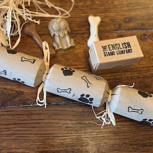 Op de afbeelding: Een verzameling items met hondenthema op een houten oppervlak. Bevat drie cilindrische crackers versierd met pootafdrukken en botten, een kleine doos van "The English Stamp Company" en hondvormig speelgoed.