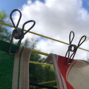May include: Two silver clothespins with spring-loaded clips are attached to a yellow clothesline. One clothespin is holding a green and white striped towel, and the other is holding a red and white striped towel.