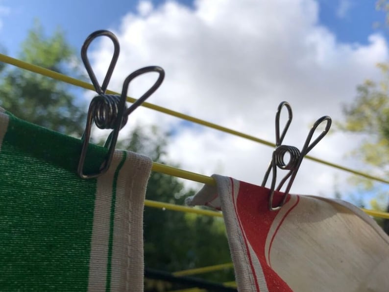 May include: Two silver clothespins with spring-loaded clips are attached to a yellow clothesline. One clothespin is holding a green and white striped towel, and the other is holding a red and white striped towel.