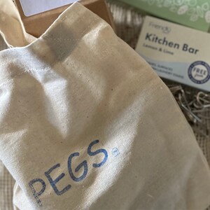 May include: A white canvas tote bag with blue lettering that says "PEGS" on the front. The bag is sitting on a brown surface.