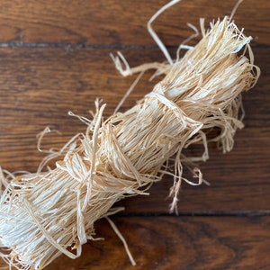 May include: A bundle of natural raffia tied with a raffia string. The raffia is a light beige color, and the bundle is resting on a dark brown wooden surface. The raffia has a textured, fibrous appearance.