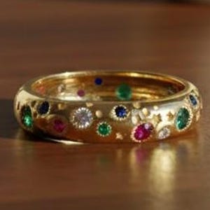Könnte beinhalten: Ein goldener Ring mit einem Band, das mit verschiedenen bunten Edelsteinen verziert ist, darunter Grün, Rot, Blau und Weiß. Der Ring hat eine polierte Oberfläche und liegt auf einer Holzoberfläche. Die Edelsteine sind in einem Muster aus Kreisen und Sternen angeordnet.