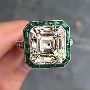 May include: A square-cut diamond ring with a green emerald border. The ring features a large, clear central stone surrounded by smaller, rectangular green stones. The setting appears to be silver or platinum.