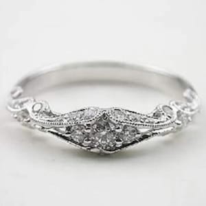 May include: A silver ring with a detailed filigree design. The ring features a central cluster of small, clear gemstones, surrounded by intricate metalwork and smaller accent stones. The band is smooth and polished.